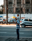 A man standing on a city street holding a phone while carrying a child on his shoulders. The background features a busy urban environment with parked cars and a partially visible building.