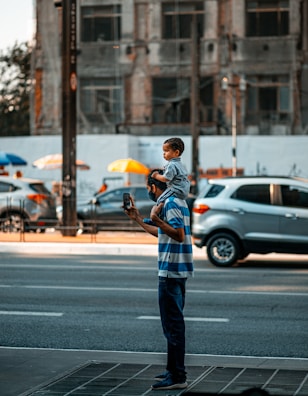 A man standing on a city street holding a phone while carrying a child on his shoulders. The background features a busy urban environment with parked cars and a partially visible building.