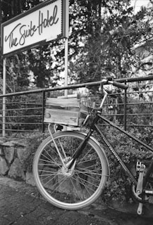 A vintage-style bicycle is parked next to a railing adorned with foliage. A sign above displays the words 'The Suite Hotel' in cursive writing. The bicycle has a wooden crate mounted on its rear rack, and the environment seems to have a rustic charm with stone paving and overgrown plants.