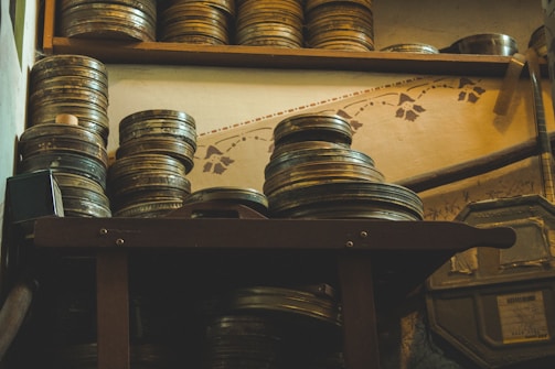 Stacks of vintage film reels are placed on a wooden shelf and table, creating a nostalgic and historical ambiance. The wall behind features decorative patterns, contributing to the vintage atmosphere.