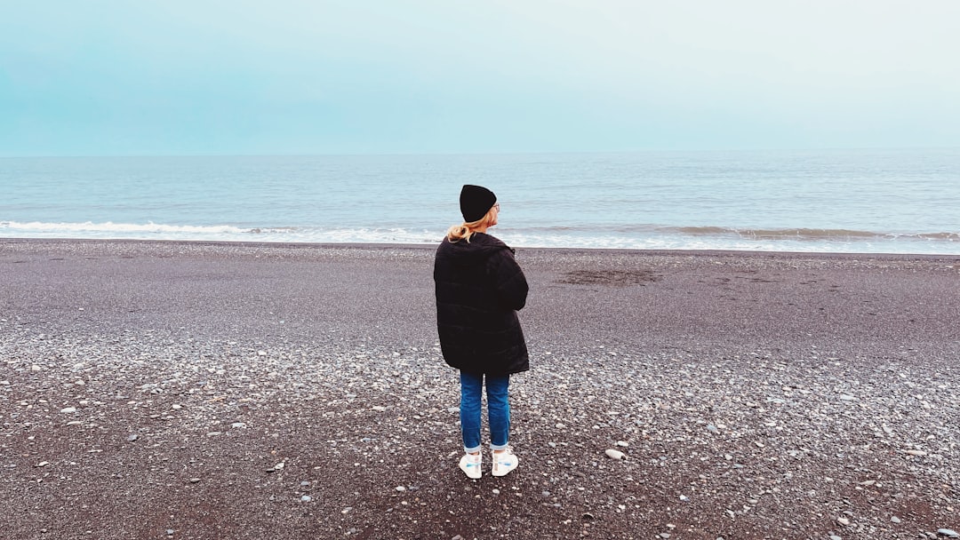 woman in black jacket and blue denim jeans standing on beach during daytime