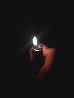 A close-up of a hand holding a lighter against a dark background. The flame is bright and provides the only source of light, illuminating the thumb and part of the lighter.