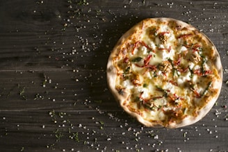 A freshly baked pizza with a crispy crust, topped with melted cheese, walnuts, sun-dried tomatoes, and fresh greens, is placed on a rustic wooden table. The table is sprinkled with salt and herbs.