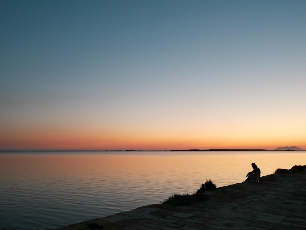 silhouette of person sitting on rock near body of water during sunset