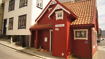 A traditional-style building painted in red with white trim is situated on a city street. The structure features a steep, tiled roof and a small front porch area. To the side, a larger, white wooden building with several windows complements the scene. The street sign reads 'Tempelkroken', and there are some plants and bags placed near the entrance.
