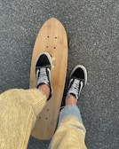 A person wearing black and white skate shoes stands on a wooden longboard skateboard on a paved surface. The person is dressed in light-colored jeans, and the pavement appears textured and slightly grainy.