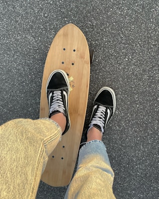 A person wearing black and white skate shoes stands on a wooden longboard skateboard on a paved surface. The person is dressed in light-colored jeans, and the pavement appears textured and slightly grainy.