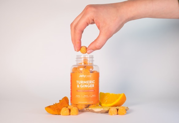 A hand is holding a gummy over a jar labeled 'Jellybee Turmeric & Ginger' that contains orange-colored gummies. Around the jar are fresh slices of turmeric root, ginger, and an orange slice, suggesting the ingredients used in the gummies. The background is a simple white, keeping the focus on the jar and ingredients.