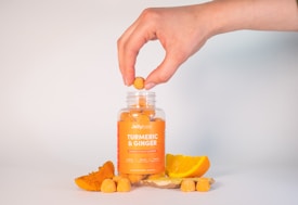 A hand is holding a gummy over a jar labeled 'Jellybee Turmeric & Ginger' that contains orange-colored gummies. Around the jar are fresh slices of turmeric root, ginger, and an orange slice, suggesting the ingredients used in the gummies. The background is a simple white, keeping the focus on the jar and ingredients.