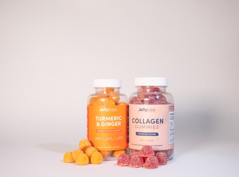 Two clear jars with white lids contain colorful gummies. The left jar is labeled 'Turmeric & Ginger' and has orange gummies, while the right jar labeled 'Collagen Gummies' holds red gummies. A few gummies are scattered in front of each jar.
