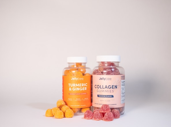 Two clear jars with white lids contain colorful gummies. The left jar is labeled 'Turmeric & Ginger' and has orange gummies, while the right jar labeled 'Collagen Gummies' holds red gummies. A few gummies are scattered in front of each jar.