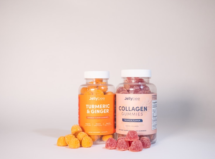 Two clear jars with white lids contain colorful gummies. The left jar is labeled 'Turmeric & Ginger' and has orange gummies, while the right jar labeled 'Collagen Gummies' holds red gummies. A few gummies are scattered in front of each jar.