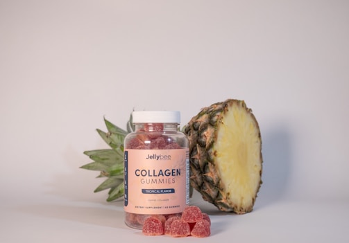 A jar of Jellybee Collagen Gummies sits next to a sliced pineapple. The gummies are tropical flavored and red in color. A few gummies are placed in front of the jar. The background is a neutral light color, which highlights the products prominently.