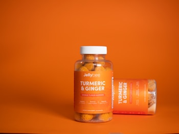 A clear plastic bottle with a white cap contains orange-colored gummies labeled as Jellybee Turmeric & Ginger. The bottle stands on an orange surface against an orange background. Next to it, another bottle lies horizontally, showing a similar label.