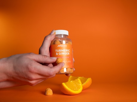 A hand holding a jar labeled 'Turmeric & Ginger Orange Flavor Gummies' against a bright orange background. In the foreground, there is a slice of orange and a gummy, both matching the vibrant hue of the setting.