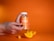 A hand holding a jar labeled 'Turmeric & Ginger Orange Flavor Gummies' against a bright orange background. In the foreground, there is a slice of orange and a gummy, both matching the vibrant hue of the setting.
