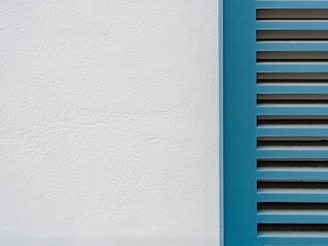 A minimalistic image featuring a close-up of a white plaster wall and a section of blue window shutters with horizontal slats.