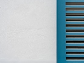 A minimalistic image featuring a close-up of a white plaster wall and a section of blue window shutters with horizontal slats.