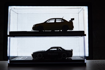 Silhouetted model cars displayed in individual glass cases are positioned against a bright backlit background, creating a sharp contrast. The upper car is a sedan with a spoiler, while the lower car has a sleek, sports-car design.