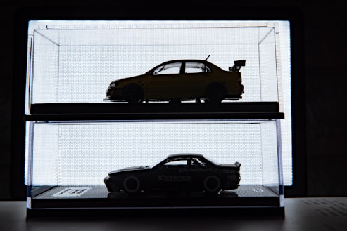 Silhouetted model cars displayed in individual glass cases are positioned against a bright backlit background, creating a sharp contrast. The upper car is a sedan with a spoiler, while the lower car has a sleek, sports-car design.