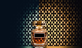 A glass perfume bottle with a golden liquid inside is placed against a patterned background. The background consists of a symmetrical floral design with a gradient from dark blue to gold. The bottle features a rectangular label and a faceted stopper, reflecting the ambient light.