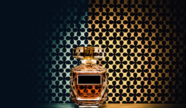 A glass perfume bottle with a golden liquid inside is placed against a patterned background. The background consists of a symmetrical floral design with a gradient from dark blue to gold. The bottle features a rectangular label and a faceted stopper, reflecting the ambient light.