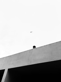 A high-contrast black and white image depicting a minimalist architectural scene. A person is partially visible, slightly peeking over a concrete ledge. In the sky above, a small airplane is seen flying. The composition emphasizes negative space and geometric forms.