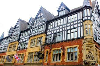 A historic building with Tudor-style architecture featuring black and white timber frames. The structure has numerous windows and pointed gables. Two flags, a Union Jack and another flag, are visible. Ornate signage for 'The Chester Grosvenor' hangs near the entrance.