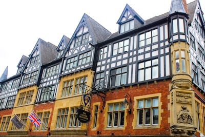 A historic building with Tudor-style architecture featuring black and white timber frames. The structure has numerous windows and pointed gables. Two flags, a Union Jack and another flag, are visible. Ornate signage for 'The Chester Grosvenor' hangs near the entrance.