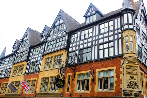 A historic building with Tudor-style architecture featuring black and white timber frames. The structure has numerous windows and pointed gables. Two flags, a Union Jack and another flag, are visible. Ornate signage for 'The Chester Grosvenor' hangs near the entrance.