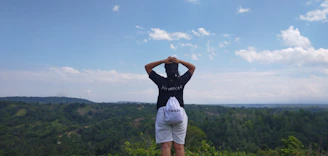 Smiling traveler standing on a hilltop overlooking lush green Uttarakhand valleys with sunlight filtering through clouds.