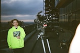 A person with wavy hair and glasses stands on a rooftop terrace holding a wine glass, wearing a neon green hoodie with a graphic print. A professional video camera on a tripod is positioned nearby, with a cityscape visible in the background under a cloudy sky. Modern buildings with glass exteriors are seen reflecting the surroundings.