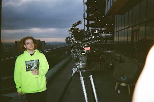A person with wavy hair and glasses stands on a rooftop terrace holding a wine glass, wearing a neon green hoodie with a graphic print. A professional video camera on a tripod is positioned nearby, with a cityscape visible in the background under a cloudy sky. Modern buildings with glass exteriors are seen reflecting the surroundings.