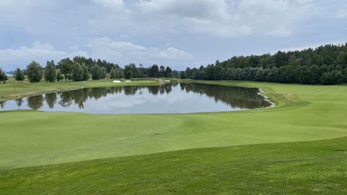 A serene landscape featuring a golf course with well-maintained green lawns and a small lake reflecting the surrounding trees and cloudy sky. Trees line the edges of the course, and the atmosphere is calm and peaceful.