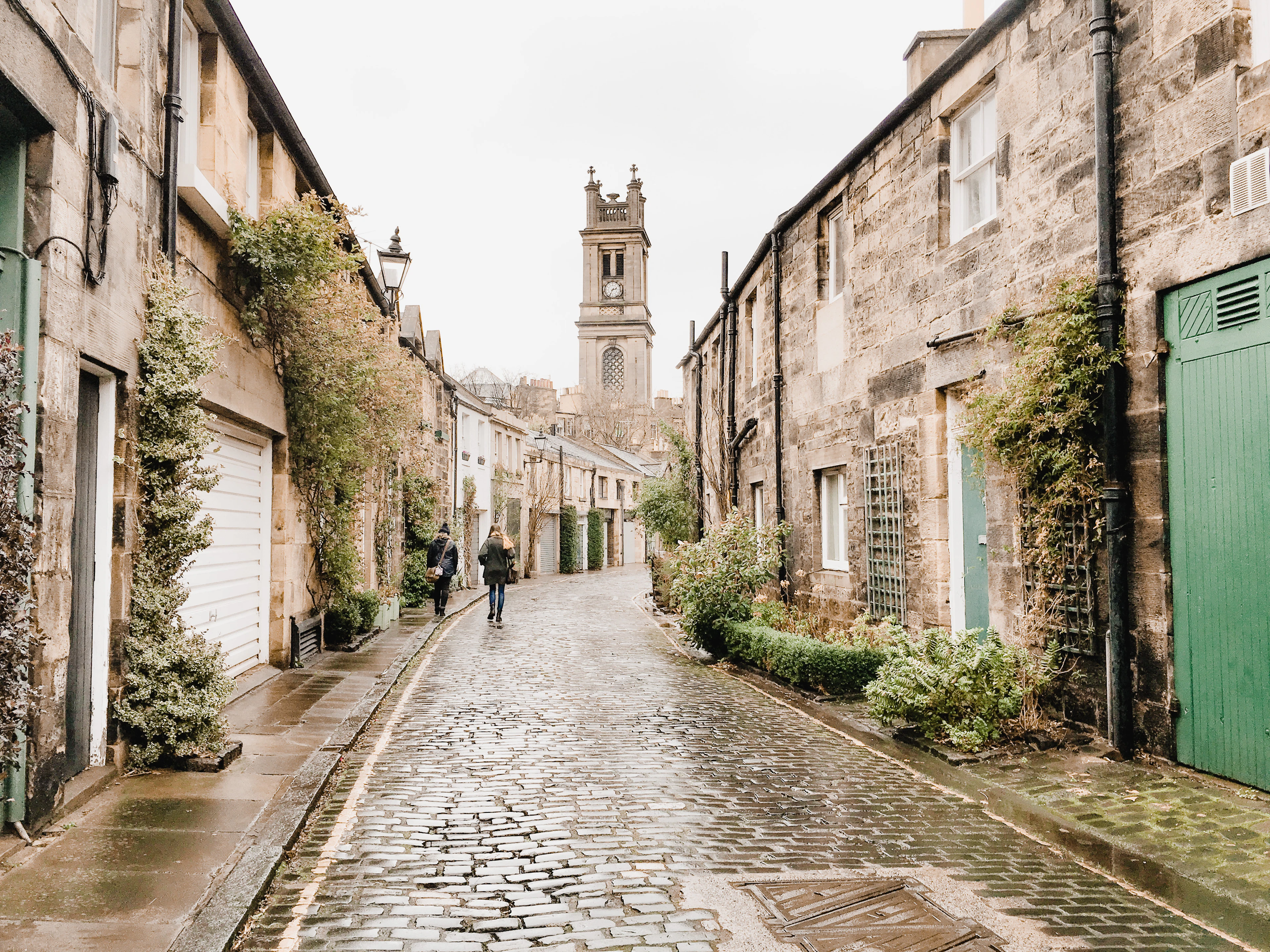 Rain-wet cobblestones on Circus Lane in Edinburgh's Stockbridge, ivy-hung mews cottages narrowing down the lane towards the clock tower of St Stephen's church at the end.