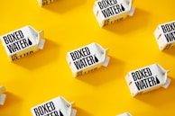 White cartons labeled 'Boxed Water Is Better' are scattered across a bright yellow background. The arrangement is neat and visually appealing, with a combination of vertical and horizontal placements creating a pattern.