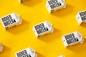White cartons labeled 'Boxed Water Is Better' are scattered across a bright yellow background. The arrangement is neat and visually appealing, with a combination of vertical and horizontal placements creating a pattern.
