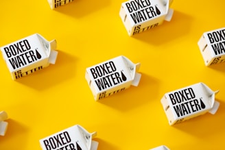 White cartons labeled 'Boxed Water Is Better' are scattered across a bright yellow background. The arrangement is neat and visually appealing, with a combination of vertical and horizontal placements creating a pattern.