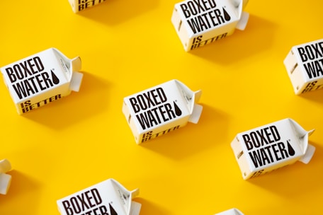 White cartons labeled 'Boxed Water Is Better' are scattered across a bright yellow background. The arrangement is neat and visually appealing, with a combination of vertical and horizontal placements creating a pattern.