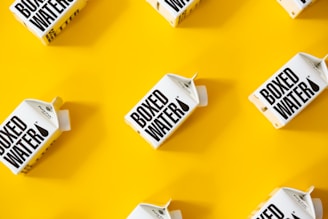 Several cartons labeled 'Boxed Water' are placed in a pattern on a bright yellow background. The boxes are white with bold black text.