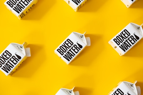 Several cartons labeled 'Boxed Water' are placed in a pattern on a bright yellow background. The boxes are white with bold black text.