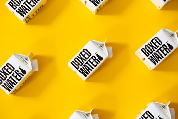 Several cartons labeled 'Boxed Water' are placed in a pattern on a bright yellow background. The boxes are white with bold black text.