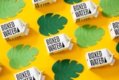 Several boxed water cartons are arranged on a bright yellow background. Green leaf cutouts are scattered around the cartons, creating a vibrant and eye-catching pattern.