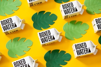 Several boxed water cartons are arranged on a bright yellow background. Green leaf cutouts are scattered around the cartons, creating a vibrant and eye-catching pattern.