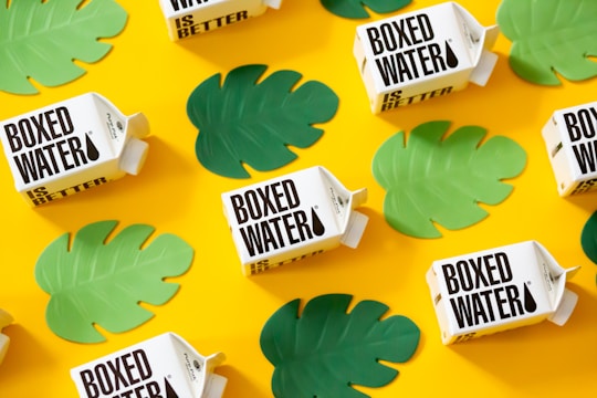 Several boxed water cartons are arranged on a bright yellow background. Green leaf cutouts are scattered around the cartons, creating a vibrant and eye-catching pattern.