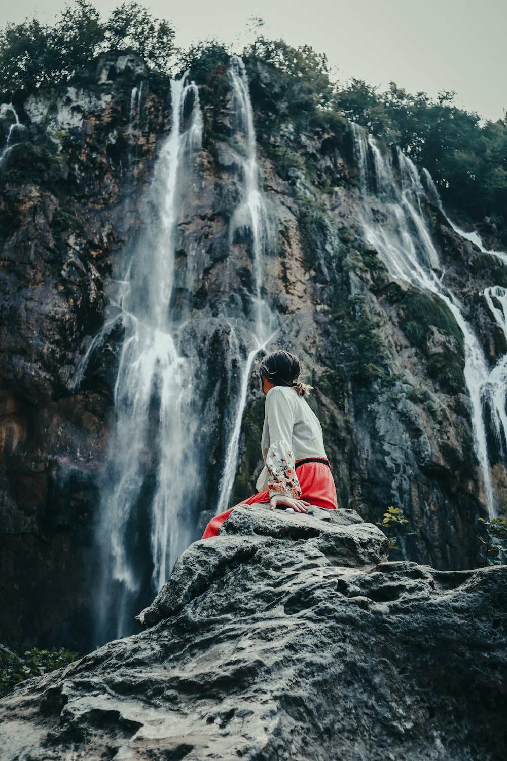 Mann in weißer Jacke sitzt tagsüber auf Felsen in der Nähe von Wasserfällen