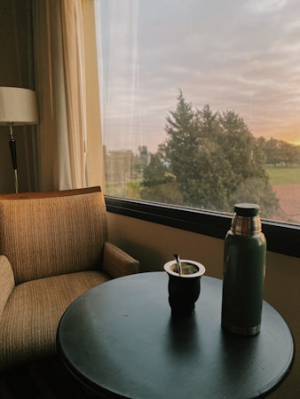 A cozy interior scene with a cushioned chair next to a round table. The table holds a thermos and a traditional mate cup with a bombilla. The background features a large window offering a view of outdoor greenery with a setting or rising sun, creating a serene and peaceful ambiance.