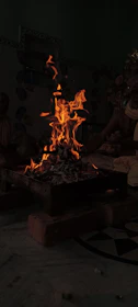A central fire pit with vibrant orange flames rising from a bed of ashes, surrounded by a darkened room. Warm hues reflect off the surrounding surfaces, creating a sense of warmth and ritual.
