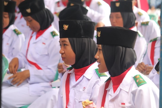 A group of young individuals dressed in white uniforms with green and red accents, wearing black hats adorned with emblems. They appear to be on a formal occasion, possibly participating in a ceremony or event.