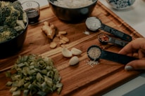 A wooden cutting board displays various fresh ingredients including chopped green onions, garlic cloves, ginger, and broccoli in a bowl. Measuring spoons hold sesame seeds, crushed red pepper, and flour. A hand is holding the spoon with sesame seeds. A small bowl with soy sauce is also visible.